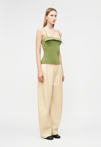 Γυναίκα που στέκεται φορώντας πράσινο αμάνικο τοπ με λεπτές τιράντες και μπεζ παντελόνι φαρδιάς γραμμής με μαύρα παπούτσια σε απλό φόντο.