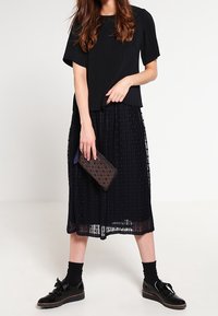 Černý vršek s plisováním a krátkými rukávy, vrstvený přes průsvitnou černou midi sukni, doplněný texturovaným černým psaníčkem a lesklými šněrovacími botami.