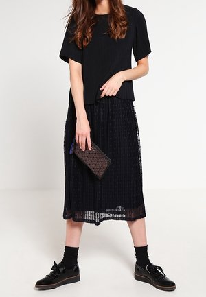 Zwarte geplooide top met korte mouwen, gedragen over een doorschijnende zwarte midi-rok, gecombineerd met een gestructureerde zwarte clutch en glanzende veter schoenen.