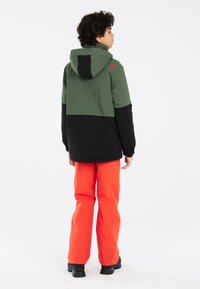 Giacca a cappuccio verde e nera con polsini elasticizzati, abbinata a pantaloni arancioni vivaci e stivali scuri, vista dal retro su uno sfondo neutro.