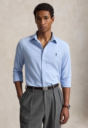 Homme portant une chemise bleu clair à manches longues avec un logo, un pantalon plissé gris, une ceinture marron et une montre-bracelet, debout avec une main dans la poche.