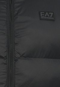 Crna puf jakna s prednjim zipom, glatke teksture i EA7 Emporio Armani logom na lijevoj strani prsnog područja.
