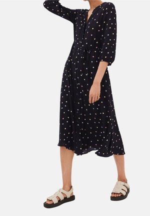 Frau trägt ein schwarzes Midikleid mit bunten Punkten, dreiviertellangen Ärmeln, Knöpfen vorne, Taillengürtel und beige offene Zehensandalen.