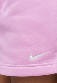 Αθλητικό σορτς σε ανοιχτό ροζ χρώμα, κατασκευασμένο από απαλό ύφασμα, με λευκό κεντητό λογότυπο Nike κοντά στο τελείωμα και ομαλή υφή.