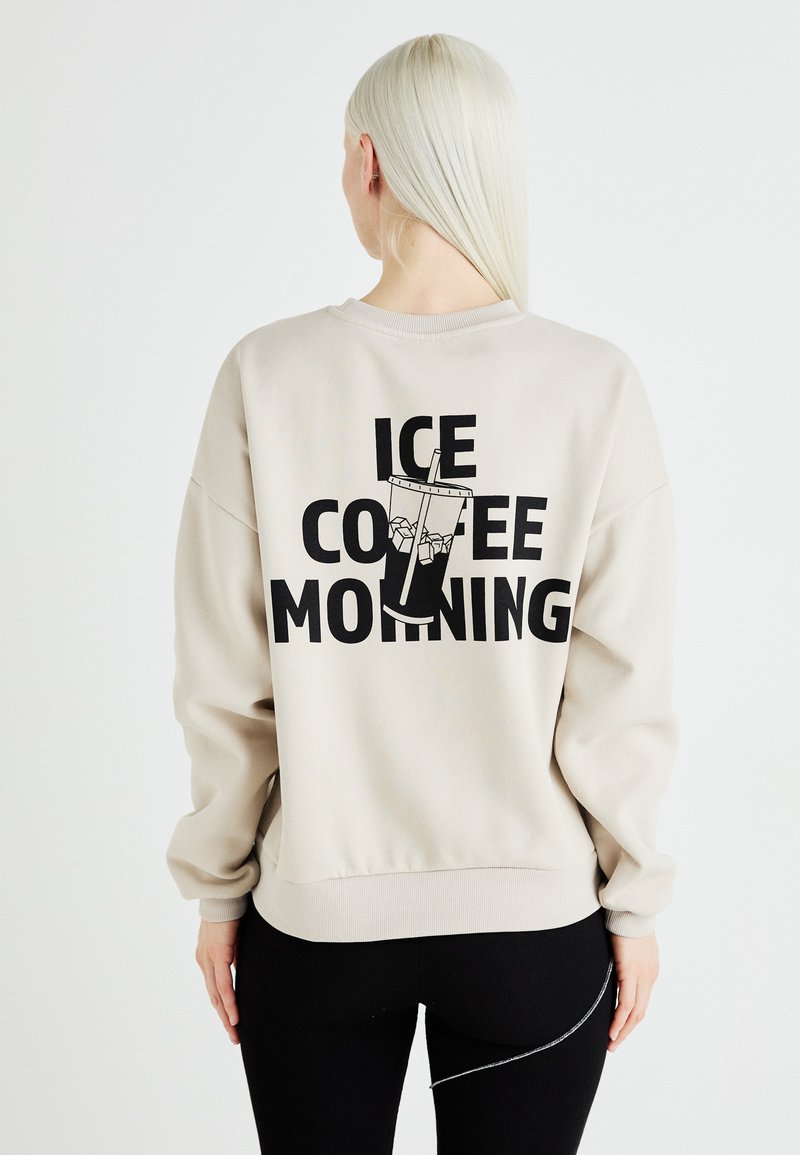 Bēšas krāsas pārāk liels džemperis ar melnu grafiku, kurā attēlots glāze ar ledu un teksts "ICE COFFEE MORNING" muguras daļā.
