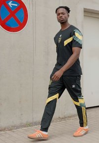 Conjunto deportivo negro con acentos en amarillo y verde, con pantalones negros y un logo de Adidas. El modelo lleva zapatillas naranjas.