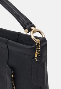 Zbliżenie na róg czarnej skórzanej torebki z wyściełanym uchwytem, złotym metalowym pierścieniem oraz czarną skórzaną metką zawieszoną na złotej łańcuszku.