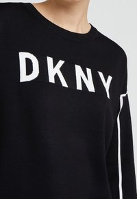 Schwarzer Strickpullover mit weißem "DKNY"-Logo. Lange Ärmel mit kontrastierendem weißen Streifen entlang der Schulter. Rundhalsausschnitt.