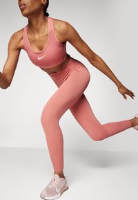 Nike sportski grudnjak i tajice od meke ružičaste tkanine, sa uskim dizajnom, glatkom teksturom i malim bijelim logotipom na grudima i gležnju.
