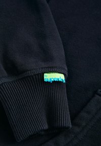 Tessuto di felpa blu navy scuro con polsini a costine. Un'etichetta brillante blu e verde con la scritta "by Superdry" è attaccata al polsino.