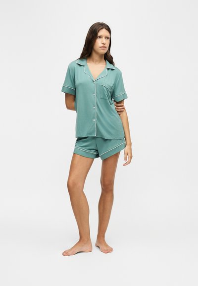 Femme debout pieds nus portant un haut turquoise à manches courtes avec boutons et un short assorti avec un liseré blanc sur un fond clair uni.