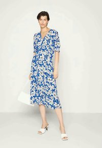 Vestido floral azul com padrões de flores brancas, mangas curtas bufantes, decote em V e comprimento midi fluido. Usado com sandálias de salto bloco brancas.