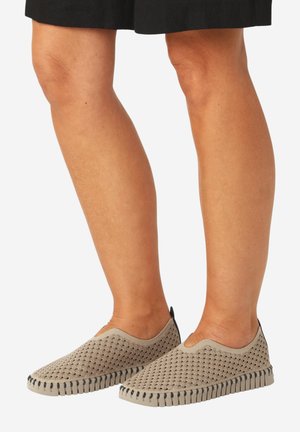 Ben iført beige slip-on sko med et bølget mønster og sorte træktabs, kombineret med sorte shorts.