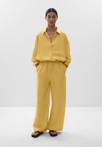 Chemise en lin jaune avec col, bouton sur le devant et poche poitrine, accompagnée d'un pantalon large assorti et de sandales noires. Texture lisse.
