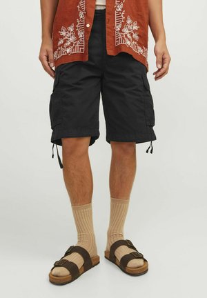 Schwarze Cargo-Shorts mit seitlichen Taschen und Zugbändern an den Säumen, kombiniert mit beigen gerippten Socken und braunen Sandalen. Eine detaillierte braun bestickte Bluse ist sichtbar.
