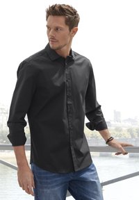 Schwarzes Hemd mit Knöpfen aus glattem Stoff, mit einem umgeschlagenen Kragen, langen, hochgekrempelten Ärmeln und einem abgerundeten Saum, kombiniert mit blauen Jeans.