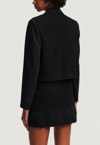 Giacca nera cropped con colletto e maniche lunghe, caratterizzata da una texture liscia e cuciture pulite; abbinata a una mini gonna a pieghe nera.