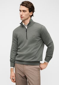 Σκούπε πράσινο πουλόβερ με γιακά μισής φερμουάρ, ριμπ μανσέτες και μικρό διακριτικό λογότυπο. Φορεμένο πάνω από λευκό πουκάμισο με γιακά, συνδυασμένο με ανοιχτό καφέ παντελόνι.