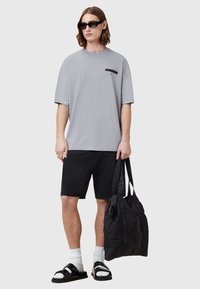 T-shirt oversize grise avec un logo devant, associée à un short noir, des sandales noires, des chaussettes blanches mi-mollet et un sac fourre-tout noir.