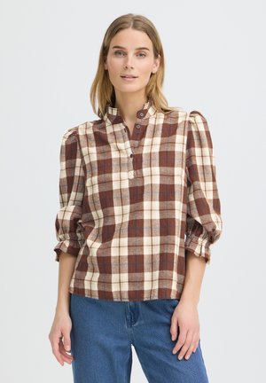 Kvinde iført brun og creme ternet langærmet bluse med rynket krave og puffede ærmer, kombineret med blå jeans på en neutral baggrund.