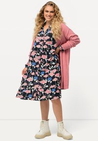 Kukkamekko, jossa on vaaleanpunaisia ja sinisiä kuvioita mustalla taustalla, yhdistettynä vaaleanpunaiseen neuleeseen ja valkoisiin platform-kengiin.