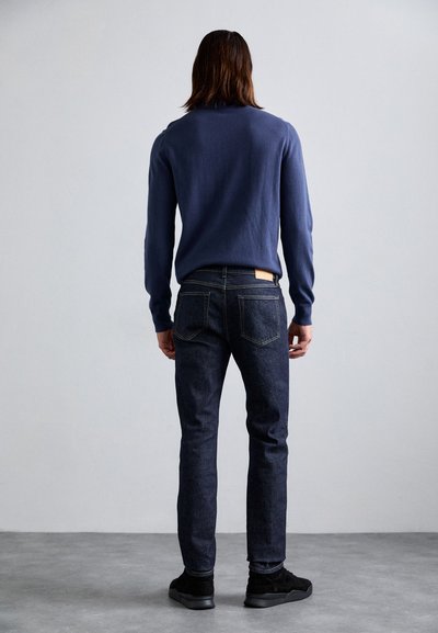 Personne avec des cheveux bruns mi-longs portant un pull marine, un jean foncé et des baskets noires, se tenant face à un mur gris uni.