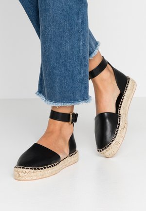 Schwarze Leder-Espadrilles mit einem Knöchelriemen, Jute-ummantelten Keilsohlen, runder Spitze und seitlichen Ausschnitten. Jeans mit ausgefranselten Säumen werden präsentiert.