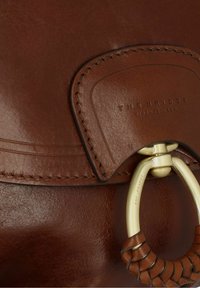Borsa a tracolla in pelle marrone con chiusura circolare in metallo dorato e un accento in pelle intrecciata testurizzata. Logo impressa sull'etichetta in pelle.