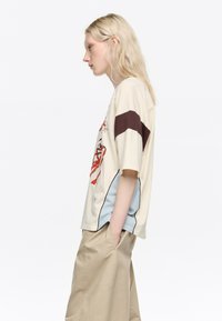 Bežna oversize majica s kratkimi rokavi, ki vključuje rdečo grafično zasnovo, rjave ramenske črte in modre stranske panele.