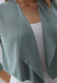 Ljus teal kardigan med öppen front, tillverkad av texturerad stickad tyg. Har trekvartsärmar och en lös, draperad silhuett.