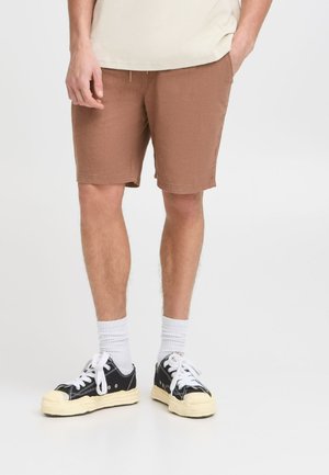 Shorts marron en tissu texturé, offrant une coupe décontractée, avec une taille élastique et des poches latérales, associés à des baskets noires et des chaussettes blanches.