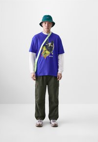 T-shirt graphique violet avec un design d'oiseau, manches longues blanches en dessous, pantalon cargo vert olive, chapeau seau vert et baskets blanches.