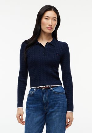 Femme aux longs cheveux foncés portant un pull polo bleu marine en maille torsadée à manches longues et un jean bleu, face à un fond blanc.