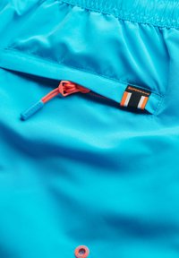 Bañador de color azul claro con un bolsillo con cremallera que tiene un tirador naranja y una pequeña etiqueta de marca en negro y naranja. Textura suave.