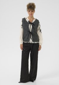 Szary wełniany kamizelka z wiązanym przodem, nałożona na kremową bluzkę z czarnym haftem. Połączona z czarnymi spodniami wide-leg w subtelne wzory.