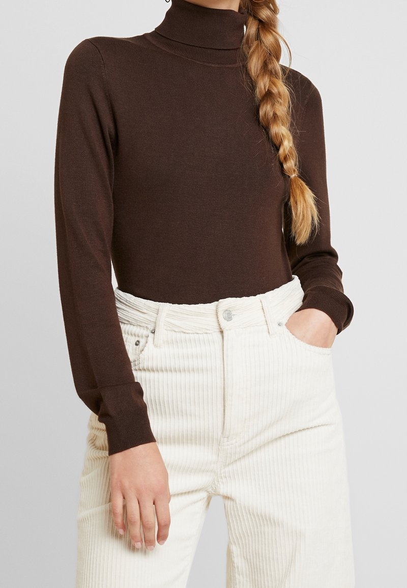 Kobieta z blond warkoczem, ubrana w brązowy sweter z wysokim kołnierzem i beżowe spodnie z sztruksu o wysokim stanie, z ręką w kieszeni na jednolitym tle.