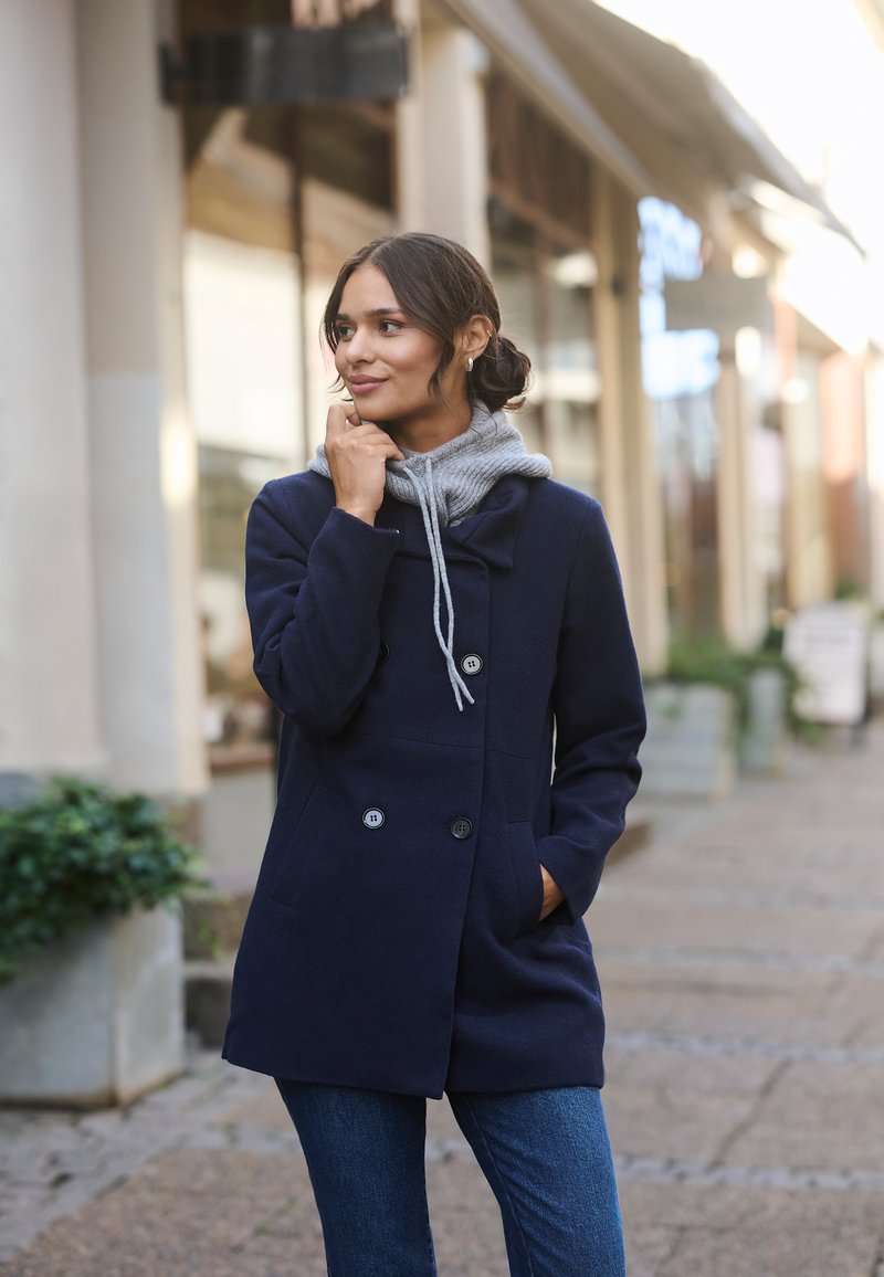 Marineblauer Wollmantel mit doppelt geknöpften Knöpfen und einer grauen Kapuzen-Innenschicht. Das Modell posiert in einer städtischen Umgebung mit blauen Jeans.