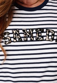 Gestreept T-shirt in marineblauw en wit, met de tekst "SAMEDI" in luipaardprint pailletten over de borst. Korte mouwen en ronde halslijn.