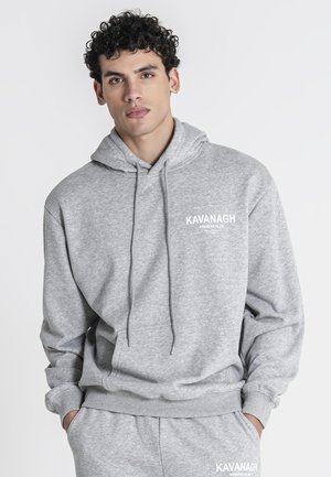 Junger Mann mit lockigem Haar, der einen hellgrauen Hoodie und passende Jogginghose trägt, beide mit weißem Schriftzug "KAVANAGH WINNERS CLUB".