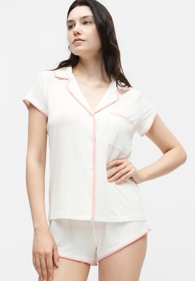 Γυναίκα με λευκό κοντομάνικο πουκάμισο με κουμπιά και αντίστοιχα σορτς με ροζ λεπτομέρειες, που ποζάρει με το ένα χέρι στη μέση μπροστά από μονόχρωμο φόντο.