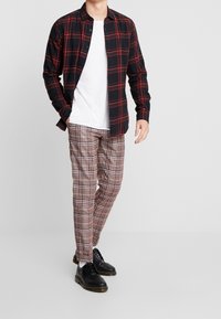 Schwarz-rot karierte Hemd, darunter ein weißes T-Shirt, braun-beige karierte Hose und schwarze Schnürschuhe. Glattes Material und lockere Passform.