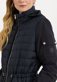 Marineblauwe donsjack met een rits aan de voorkant, een capuchon, elastische manchetten en een getailleerde taille. Bevat een patchzak en logo-detail op de mouw.