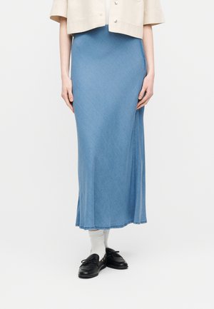 Femme portant une jupe bleu clair longueur cheville, une veste courte beige, des chaussettes blanches et des mocassins noirs, debout devant un fond uni.