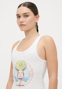 Camiseta sin mangas blanca de tejido acanalado, con un gráfico colorido de una mujer con una rodaja de lima en la cabeza y el texto "Está en lo ácido".