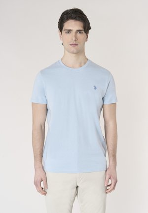 Giovane uomo con capelli scuri che indossa una maglietta a maniche corte azzurro chiaro e pantaloni beige, in piedi davanti a uno sfondo semplice.