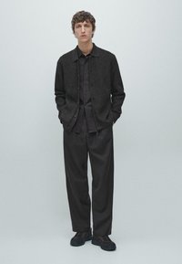 Junger Mann steht mit den Händen in den Taschen, trägt eine schwarze, gerippte Strickjacke, ein schwarzes Hemd mit Knöpfen, lockere schwarze Hosen und schwarze Sneakers.