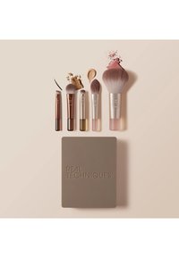 Fem sminkebørster merket RT med prøver av foundation, pudder og blush over, plassert over en oppbevaringsetui fra Real Techniques.