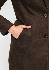 Manteau en mélange de laine marron au fit ajusté, avec une poche latérale et une fermeture à bouton, mettant en valeur une finition extérieure lisse et texturée.