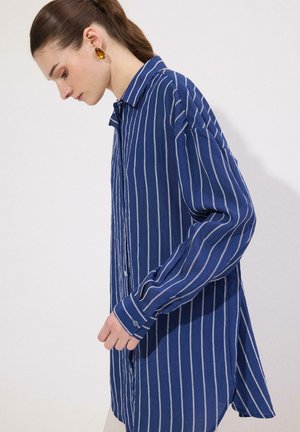 Γυναίκα με καστανά μαλλιά που φοράει κεχριμπαρένια σκουλαρίκια και ένα άνετο μπλε πουκάμισο με λευκές κάθετες ρίγες, στραμμένη προς τα αριστερά σε απλό φόντο.