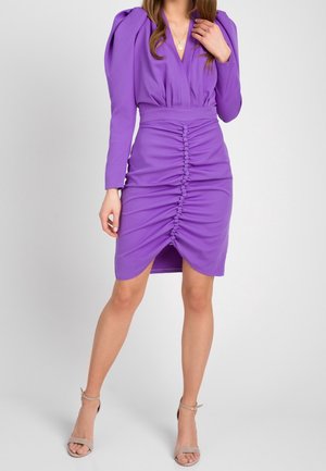 Kobieta nosząca dopasowaną fioletową sukienkę z długimi rękawami, marszczony przód z guzikami obciągniętymi tkaniną, i beżowe szpilki na pasku wokół kostki, stoi na szarej podłodze.